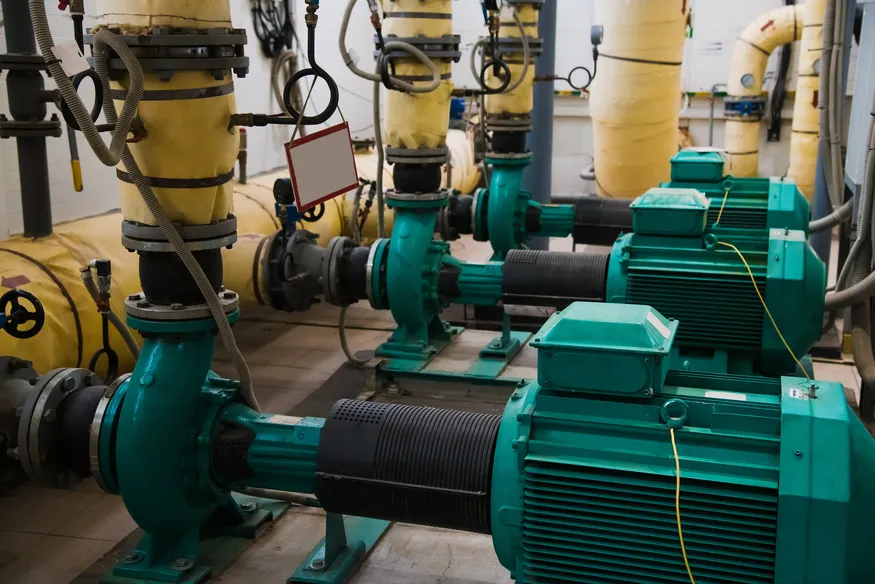 Fila de bombas de agua industrial conectadas a tuberías amarillas aisladas térmicamente en un cuarto de máquinas cerrado.