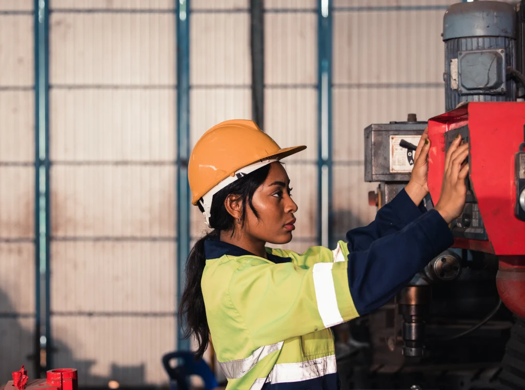 Una técnica en overol amarillo y casco naranja opera una máquina industrial, un ejemplo de mantenimiento correctivo.